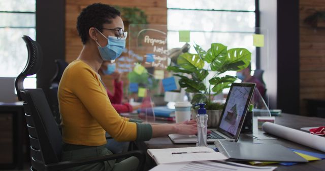 Young woman working on a laptop in a modern office, with team members visible in the background. Wearing a face mask, the professional is adhering to health and safety precautions. This can be used for illustrating modern work environments, office culture during pandemic, remote collaboration setups, and health safety adherence in professional settings.