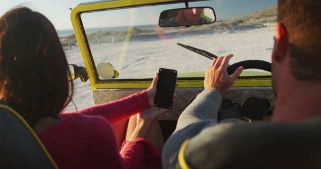 Happy caucasian couple sitting in beach buggy by the sea using smartphone. beach stop off on summer holiday road trip.