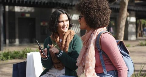 Young women sharing smartphone and chatting on sunlit city bench after shopping