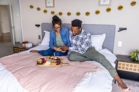 Couple exchanging ring on cozy bed with charcuterie board and romantic bedroom decor