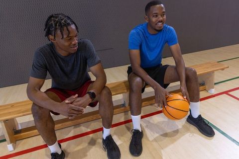 Two african american friends sitting on bench holding basketball in indoor gymnasium