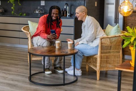 Diverse friends laughing and sharing smartphone in cozy coworking lounge with coffee