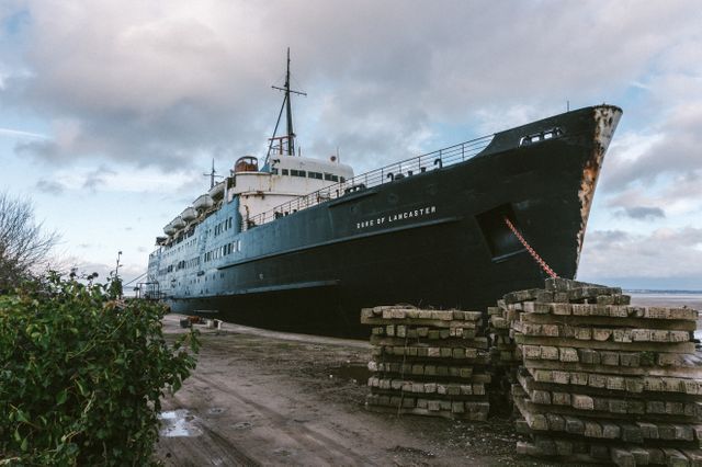 This image of a historic cruise ship named 'Duke of Lancaster' moored at an abandoned dock can be used to convey themes of past maritime glory and industrial decline. The overcast sky adds to the melancholic atmosphere, making it suitable for projects focusing on abandoned industrial sites, historic preservation, and outdoor adventures.