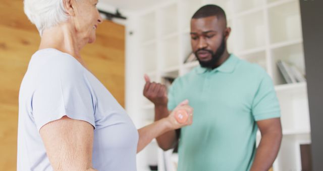 This shows a senior woman receiving guidance from a trainer while exercising with a dumbbell inside a home environment. It is good for illustrating physical therapy, health, fitness, elderly care, and wellness themes.