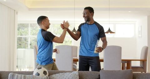 Diverse friends high-fiving while celebrating soccer win in sunlit modern living room