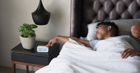Reaching for alarm clock in morning, young man waking under white duvet in modern bedroom