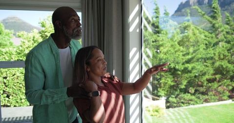 Couple pointing toward backyard while checking smartwatch in bright home with mountain view