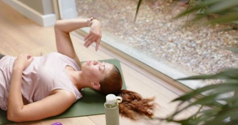 Young woman relaxing on yoga mat pointing during post-workout cooldown by sliding door