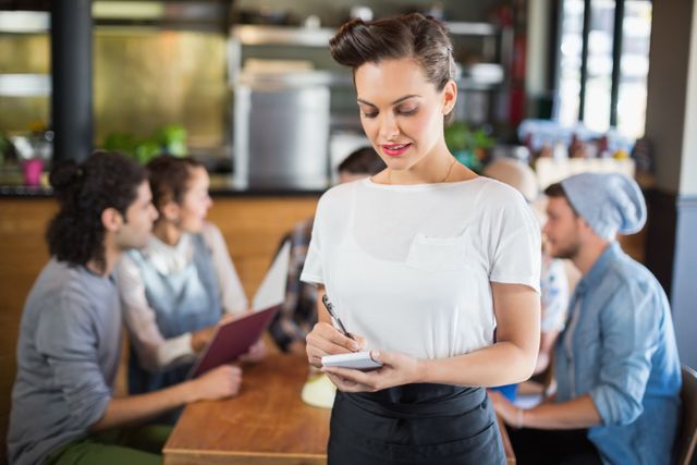 Beautiful waitress writing while standing by customers in restaurant