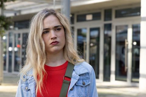Young woman standing outside glass doors looking thoughtful in denim jacket and red top