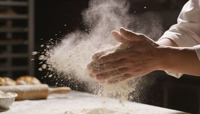Baker clapping floured hands creating dramatic flour cloud over wooden workbench