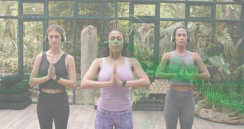 Three women meditating in greenhouse wearing headphones and workout clothes for wellness