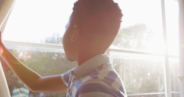 African american boy opening curtain and looking out of sunny window, backlit by sun. spending free time at home.