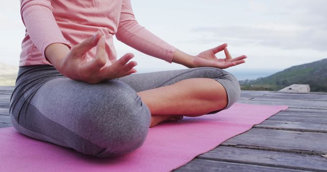 Individual sitting in lotus position on pink yoga mat practicing meditation on wooden deck with scenic background. Ideal for topics related to wellness, mindfulness, outdoor exercise, relaxation techniques, and healthy lifestyle promotion.