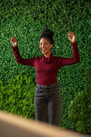 Joyful african american woman raising arms wearing burgundy sweater against green wall
