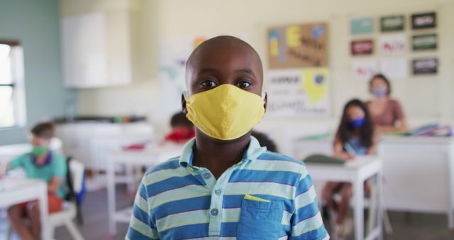 Boy in yellow face mask standing in classroom with other masked students in background. Useful for illustrating school safety protocols, education during pandemic, children's health, and classroom settings.