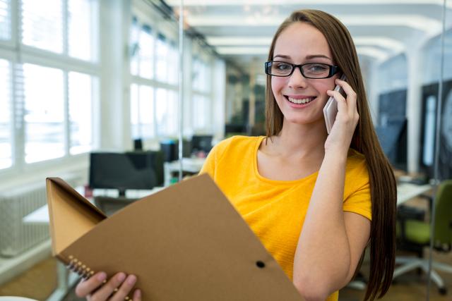 Young businesswoman holding a file and talking on the phone in a modern office. Ideal for use in corporate websites, business presentations, marketing materials, and articles about professional communication and office environments.