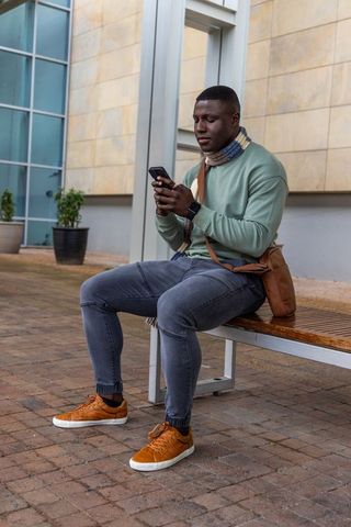 African american man checking smartphone while sitting on urban bench wearing knit scarf