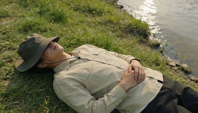 Man resting on grassy lakeside wearing olive hat and khaki shirt with wristwatch