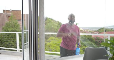 African american man cleaning balcony glass using blue sprayer overlooking suburban terrace