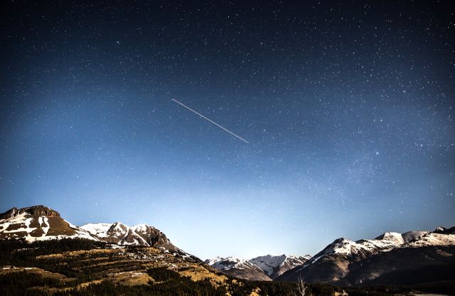 Night sky filled with stars above snow-capped mountains, highlighting a meteor streak across the sky. Ideal for projects related to nature, nightscapes, stargazing, astronomy, wilderness adventures, and outdoor serenity concepts.
