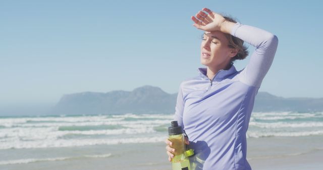 The image shows a woman holding a water bottle and shielding her eyes from the sun, taking a break from running on the beach. This image can be used for content related to fitness, outdoor activities, hydration, healthy living, and summer exercise routines.