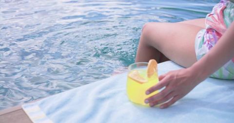 Woman sitting poolside dipping foot while holding citrus drink on towel for summer relaxation