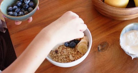 Placing blueberries on granola and banana bowl on wooden table with yogurt and citrus