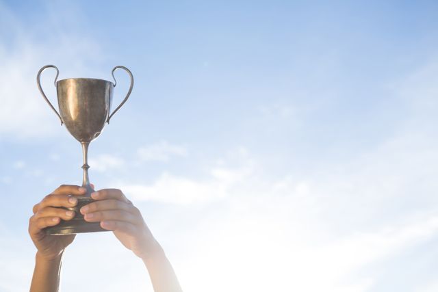 Hands holding a trophy high against a blue sky with clouds. Ideal for concepts of achievement, success, victory, and celebration. Perfect for use in motivational materials, award ceremonies, and recognition events.