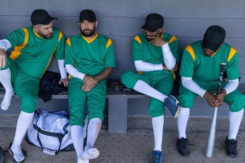 Diverse baseball teammates sitting on dugout bench wearing green uniforms holding bat