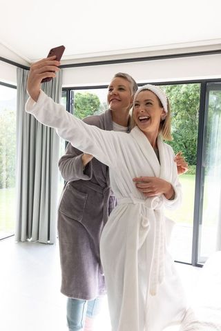 Two women wearing plush bathrobes taking joyful selfie in modern bedroom with garden view