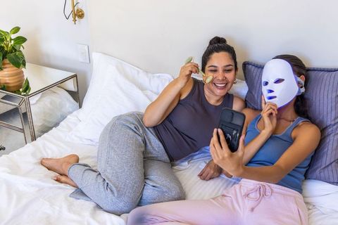 Multicultural friends relaxing with jade roller and led face mask taking selfie in bed