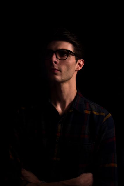 This image shows a thoughtful young man wearing glasses and a plaid shirt, standing in a dark room with intense, focused lighting highlighting his profile. The mysterious and contemplative atmosphere makes this image suitable for use in publications and projects focusing on introspection, isolation, or dramatic themes. It could also be used for psychological content, or in fashion campaigns highlighting casual outfits and eyewear.