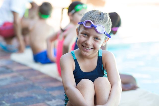Boy sitting at the edge of a pool, wearing swimwear and goggles, smiling at the camera. Other children in the background, also in swimwear, suggesting a group swimming activity. Ideal for use in advertisements for swimming lessons, summer camps, or children's activities.