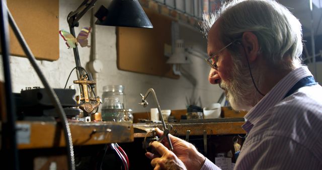 Elderly man concentrating on intricate work at workshop desk, surrounded by tools and materials. He wears a beard and focuses intently on his craft. Suitable for themes related to dedicated craftsmanship, occupational skills, and senior hobbies. Can be used in articles about lifelong learning, features on artisans, or crafting tutorials.