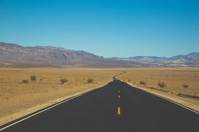 This image shows an empty desert highway stretching towards distant mountains under a clear blue sky. It is perfect for themes of travel, adventure, and road trips. Suitable for blogs, travel agencies, or inspiration boards highlighting the beauty of remote and untouched landscapes.