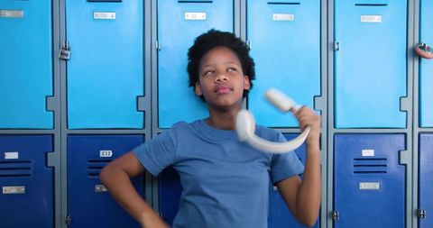 Teen student leaning on blue lockers holding white headphones wearing blue tee looking thoughtful