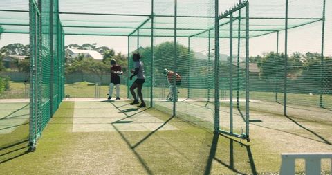 African american coach bowling to indian batter practicing batting in outdoor cricket nets