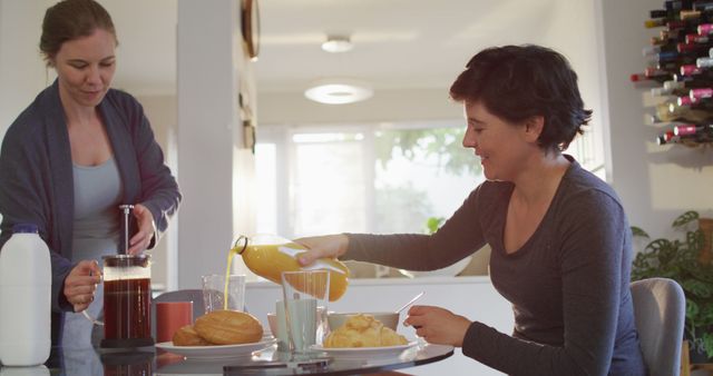 Two women in casual clothing preparing breakfast at home in the morning. One is pouring orange juice while the other is making coffee. Various breakfast items like bread, pastries, and a milk container sit on the table. Light-filled kitchen with modern decor visible. Suitable for concepts related to healthy breakfast, domestic life, morning routine, friendly interaction, and home cooking.