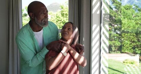 Mature couple sharing tender embrace by sunlit window, smartwatch, home intimacy, afternoon light