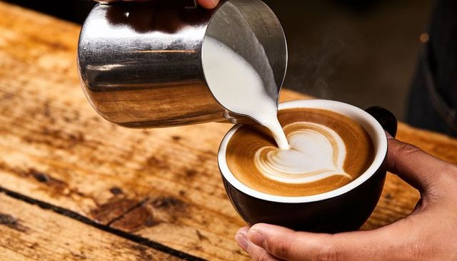 Barista pouring heart latte art into black cup on rustic wood counter