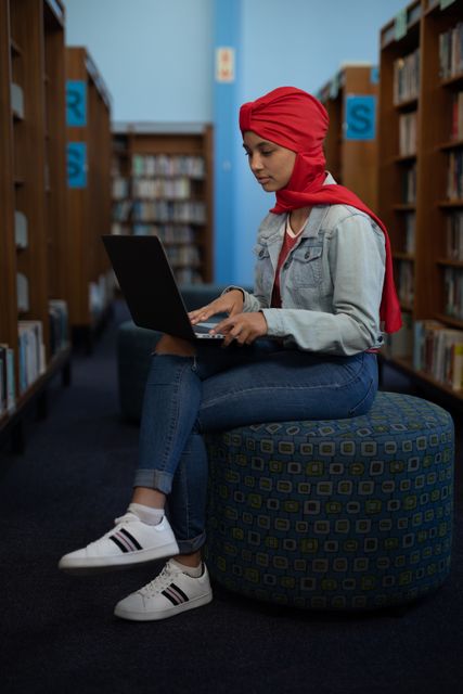 Side view of an Asian female student wearing denim jacket studying in a library sitting on a seat between the bookshelves using a laptop computer.