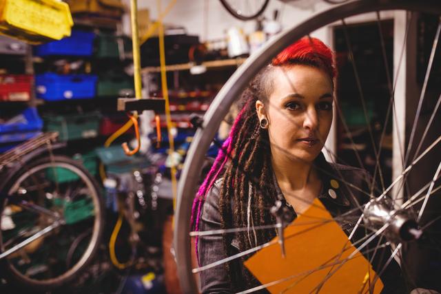 Female mechanic with dreadlocks and punk style examining a bicycle wheel in a workshop filled with tools and equipment. Ideal for use in articles or advertisements related to bicycle repair, mechanics, women in trades, and industrial work environments.