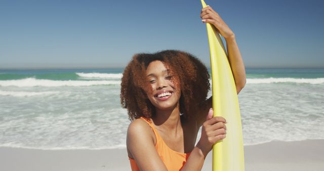 Biracial woman with curly hair standing on beach with surfboard and laughing. Summer, vacation, relaxation, happy time.