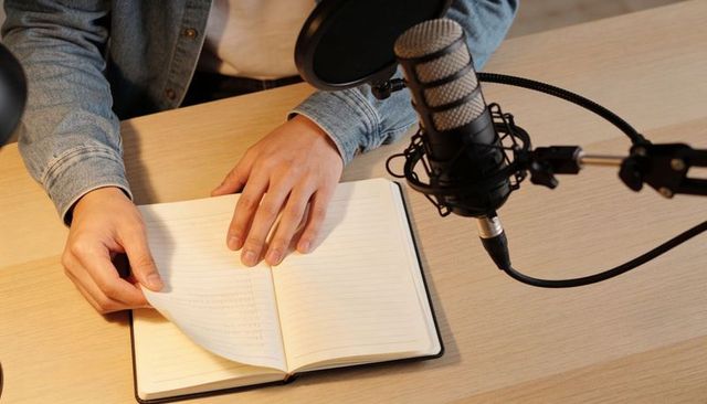 Podcast host preparing notebook at microphone with pop filter on light wood desk