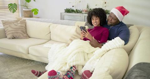 Couple cuddling on sofa under chunky blanket, scrolling smartphone, wearing Santa hat