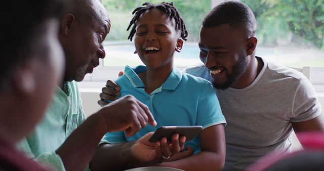 Grandfather, father, and son gathered at home, sharing a moment of laughter while looking at a smartphone. Relatable and heartwarming scene illustrating familial love and enjoyment. Perfect for materials focusing on family bonding, technology in everyday life, and multigenerational relationships.