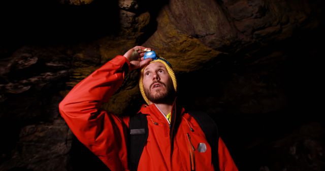 Man wearing red jacket and yellow beanie exploring a dark cave with a headlamp, looking up towards rock walls. Ideal for use in articles or advertisements about outdoor adventures, cave exploration, travel destinations, or spelunking equipment. Perfect for travel blogs, adventure magazines, and outdoor gear promotions.