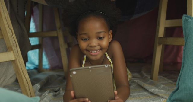 Young African American girl smiling while using a tablet inside a homemade play fort. Ideal for themes involving childhood, technology, education, and early learning. Perfect for family-oriented advertisements, e-learning platforms, or content promoting imaginative play and home activities for children.