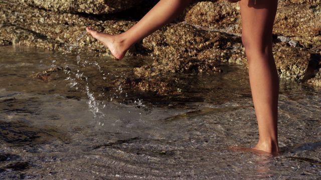 Barefoot woman enjoying sunny day on rocky beach, playfully splashing water with her leg. Ideal for themes related to beach vacations, summer fun, outdoor activities, and relaxation by the ocean.